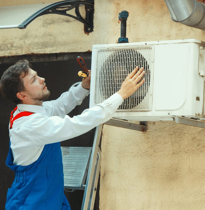 HVAC technician on a ladder repairing a wall-mounted outdoor air conditioning unit using hand tools.